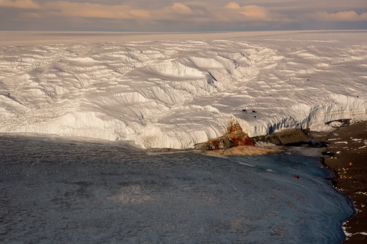 Panoramic view of the McMurdo Dry Valleys in Antarctica, showing Blood Falls as a red streak against the white Taylor Glacier and surrounding barren landscape.