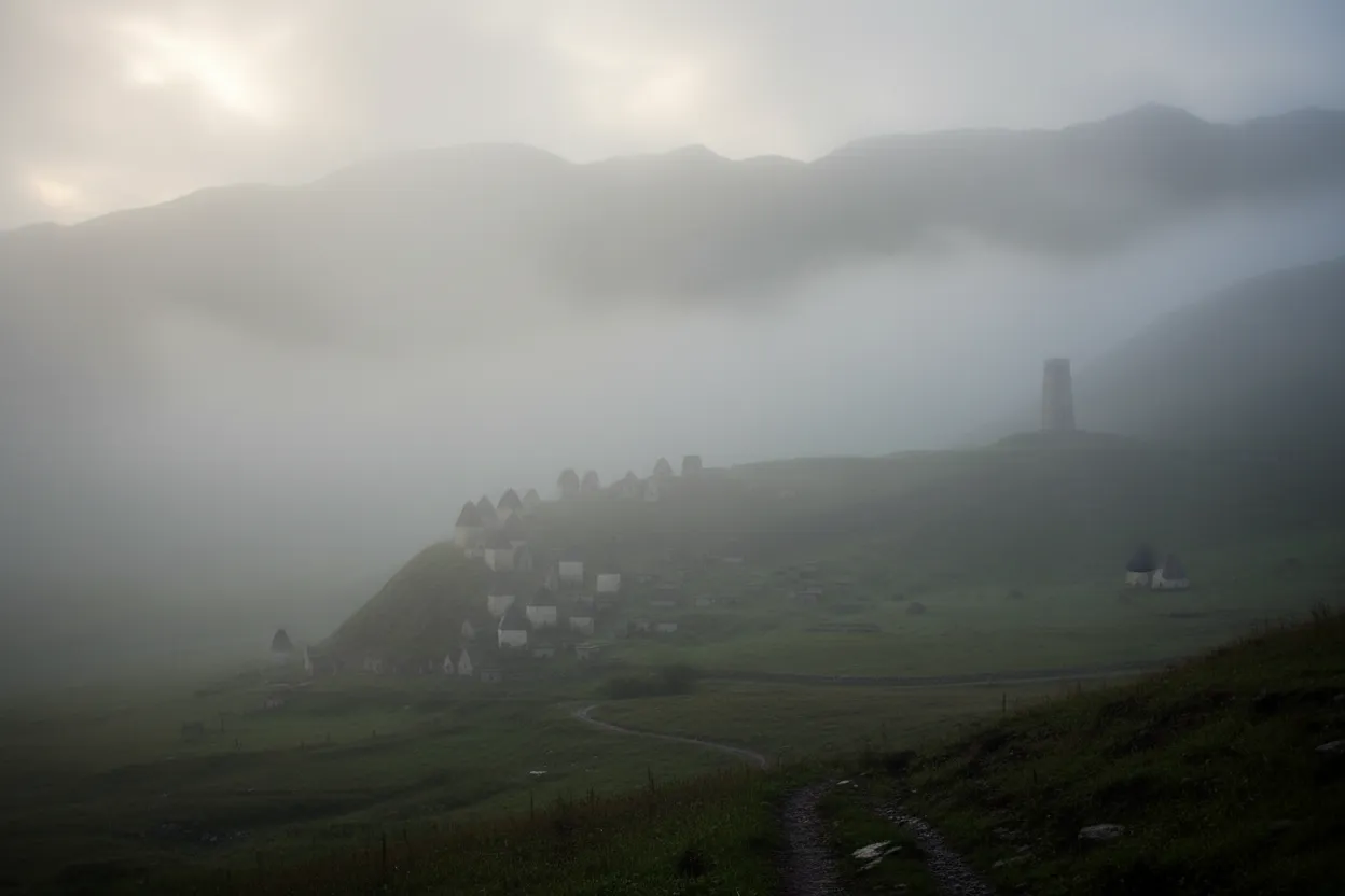 A winding mountain road leading through a lush green valley with towering peaks, hinting at the journey to Dargavs, with a small village visible in the distance.