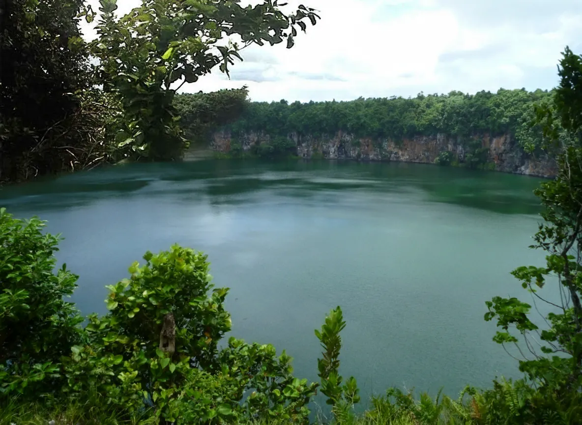 Lush green tropical rainforest bordering the serene waters of Lake Lalolalo, with light filtering through the canopy.