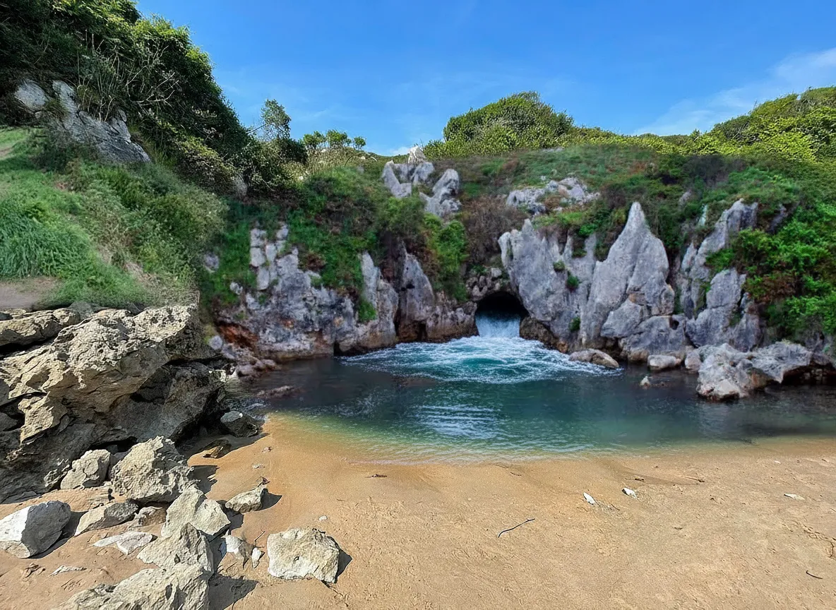 Playa de Gulpiyuri, a small crescent-shaped beach surrounded by green grass, with clear blue water and gentle waves.