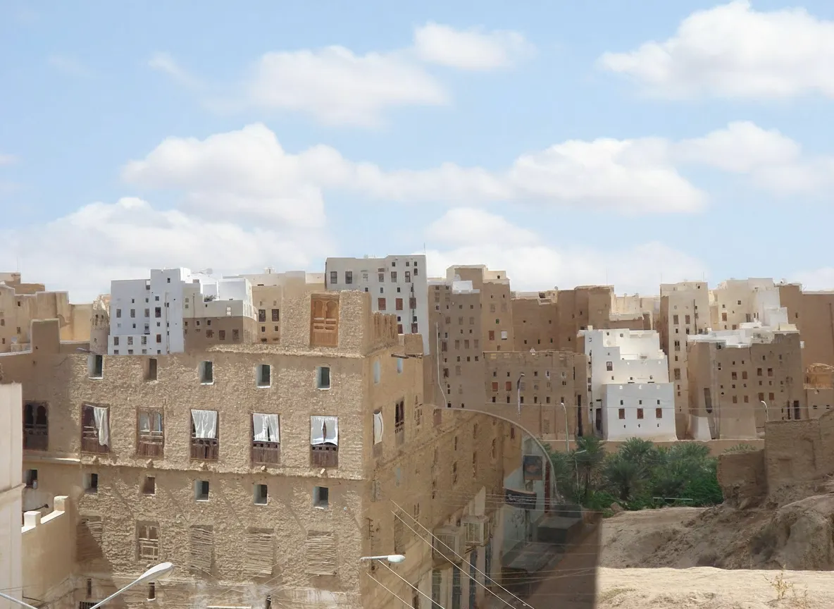 Narrow, winding alleyway in Shibam, Yemen, flanked by towering mud-brick buildings with intricate wooden doors and windows.