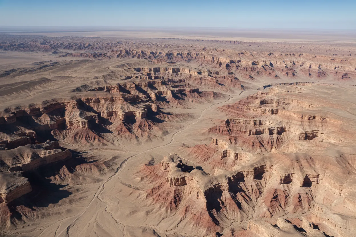 A lone figure stands at the edge of Yangikala Canyon, providing a sense of scale against the massive, colorful rock formations, bathed in warm afternoon light.